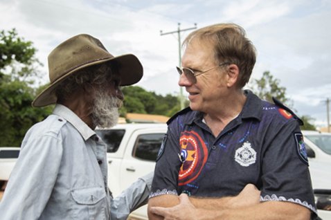 Photo of a person in an indigenous artwork QPS shirt talking to another person.