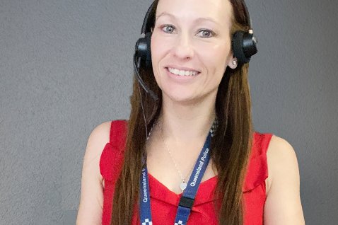 Photo of a person wearing a stereo headset with microphone, a red top and a Queensland Police Service lanyard. The background is a blank grey wall.