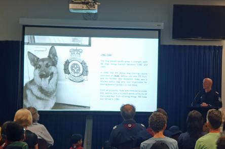This is an image of an auditorium with people sitting on chairs facing a projector screen and a police officer who is giving a talk about the history of the dog squad. The projector screen is showing a german shepard dog, named Duke, sitting next to a police car. The image was probably taken sometime in the mid 1980's.