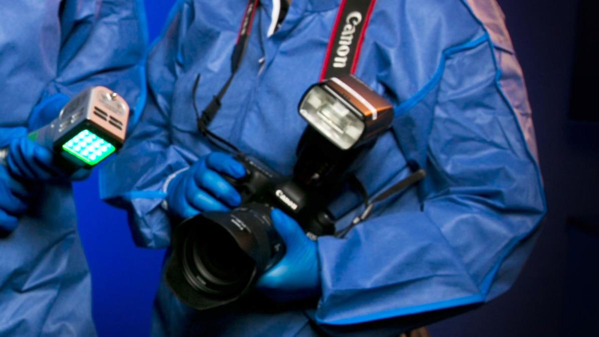 Forensic officer in blue coveralls and gloves, holding photographic equipment