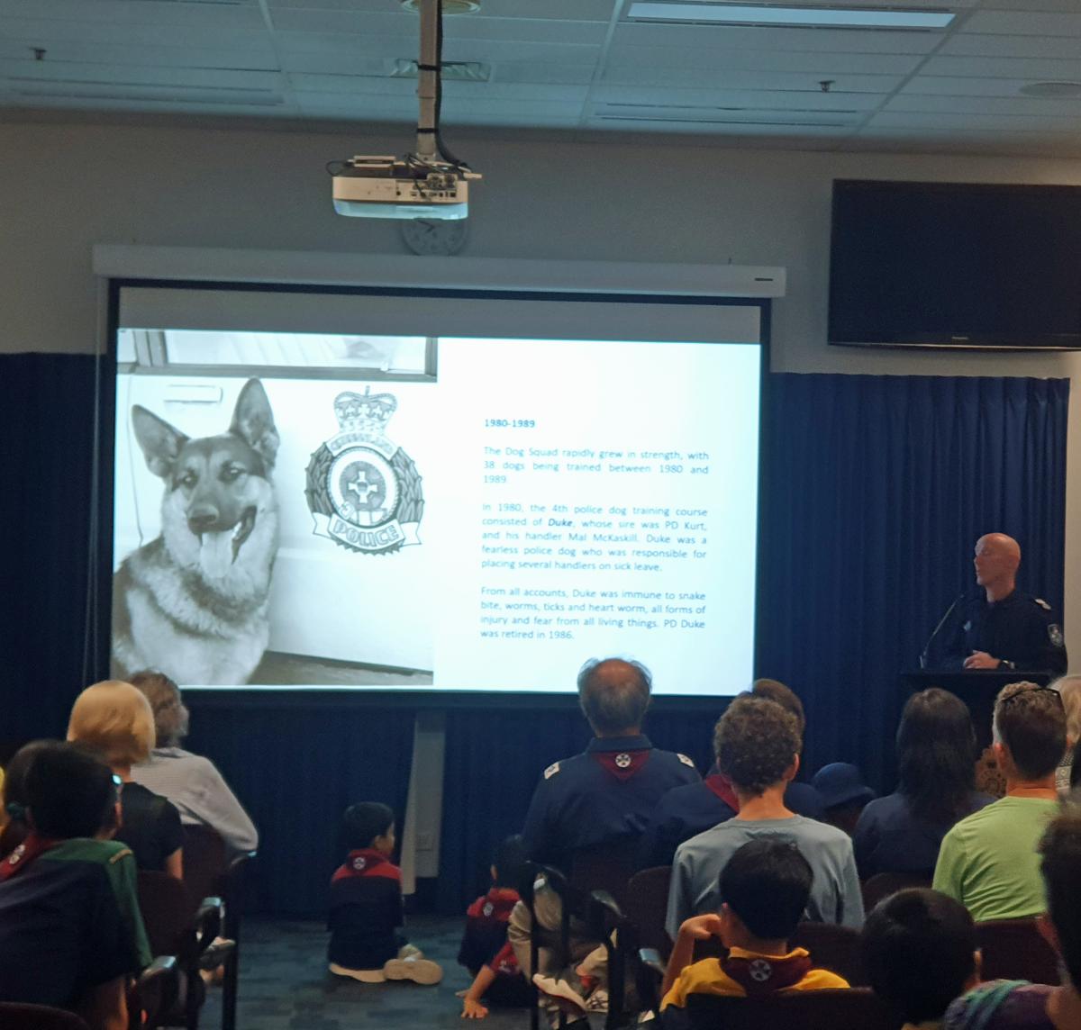 This is an image of an auditorium with people sitting on chairs facing a projector screen and a police officer who is giving a talk about the history of the dog squad. The projector screen is showing a german shepard dog, named Duke, sitting next to a police car. The image was probably taken sometime in the mid 1980's.